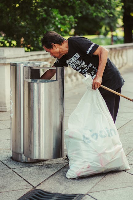 Three large black garbage bags filled with waste are placed on the pavement beside a black metal fence, which features vertical bars and a lattice pattern at the top. The bags appear to be made of thi